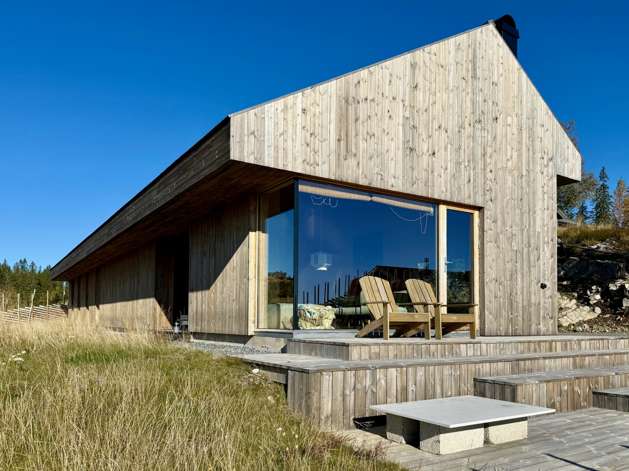 Nowoczesny dom z drewna o minimalistycznej architekturze, z dużym przeszkleniem i drewnianym tarasem. Widoczne krzesła na tarasie i trawa na pierwszym planie.