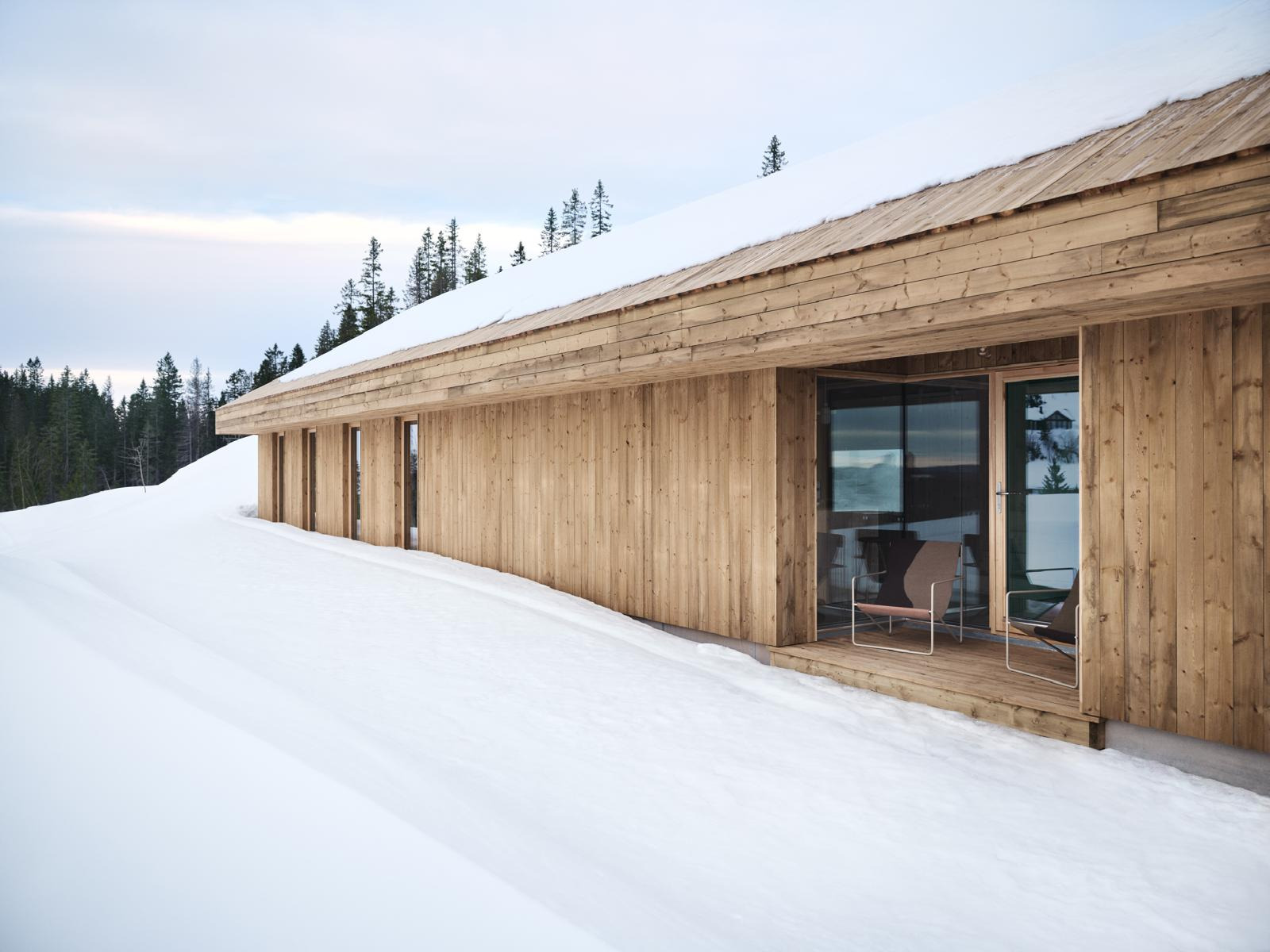 Drewniany dom w zimowym krajobrazie, zadaszony taras z krzesłami, minimalistyczny design, naturalne oświetlenie, widok na las w tle, śnieżny teren.
