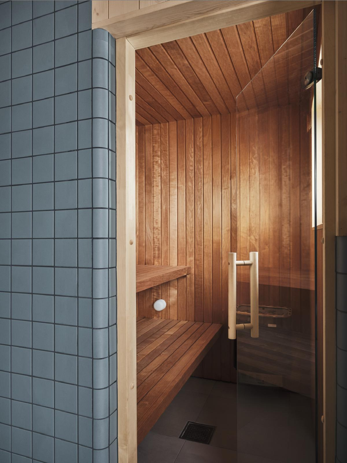 Nowoczesna sauna z drewnianym wnętrzem i szklanymi drzwiami, wykończona szarymi płytkami. Widoczne drewniane ławki i oświetlenie. Minimalistyczny design.