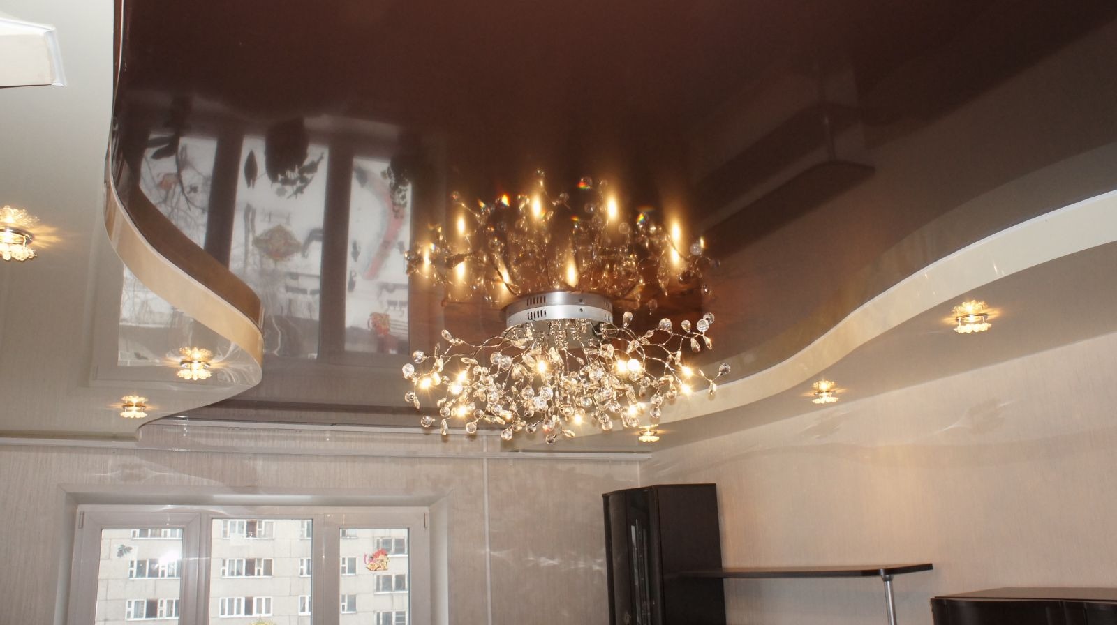 Dwupoziomowy, brązowo-biały sufit napinany z połyskiem, oświetlony dekoracyjnym żyrandolem i małymi lampkami. Widoczne okno z odbiciem w suficie.