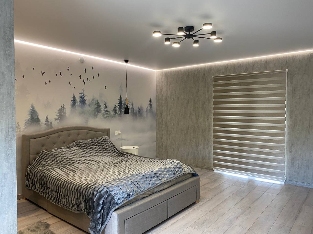 Nowoczesna sypialnia z łóżkiem, tapetą z motywem lasu i ptaków, oświetleniem LED podsufitowym i roletami okiennymi w neutralnych barwach.
