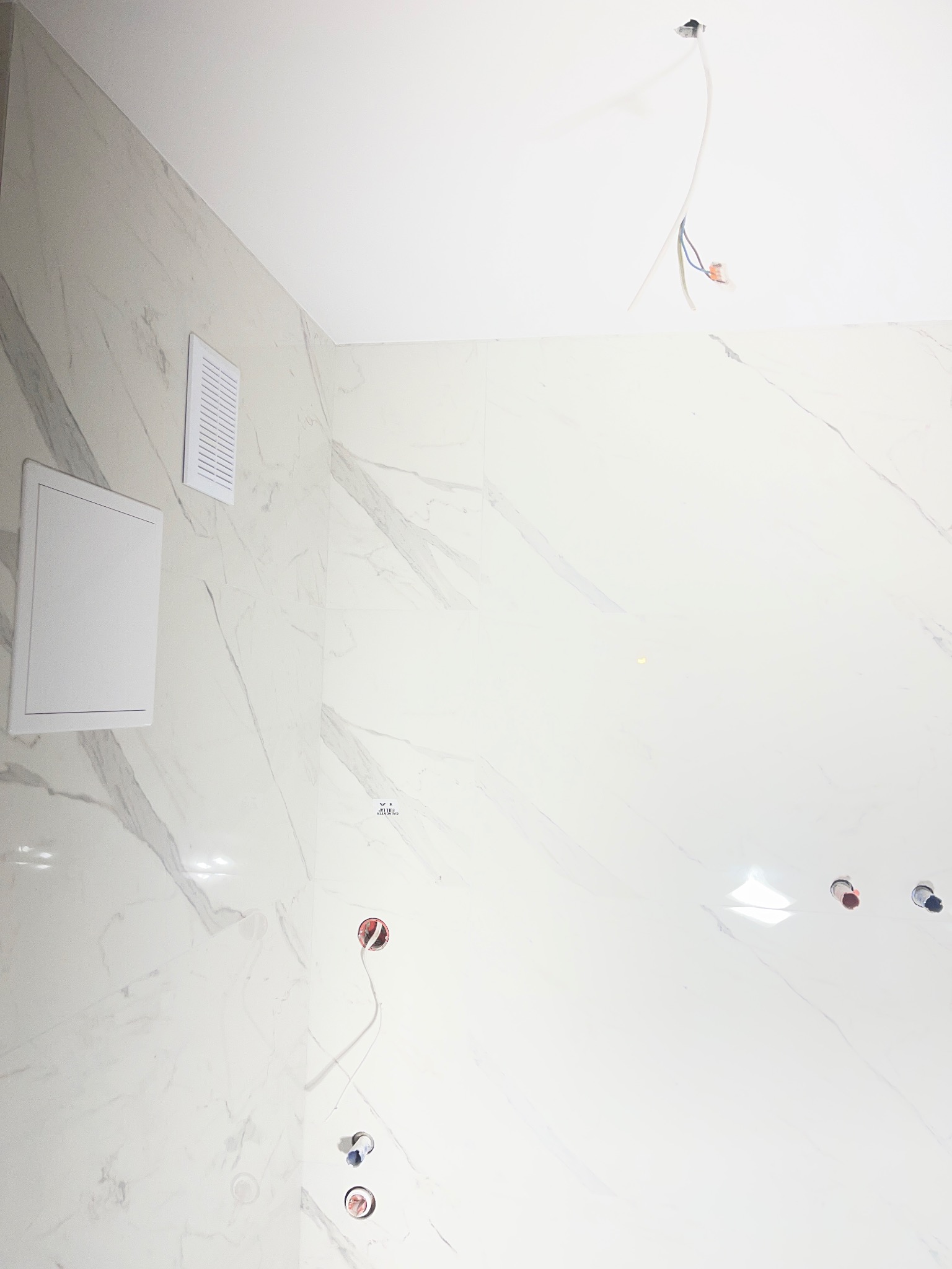 Wykończona łazienka z białymi płytkami imitującymi marmur, widoczne otwory na armaturę i wystające przewody elektryczne. Zamontowany wentylator i rewizja.