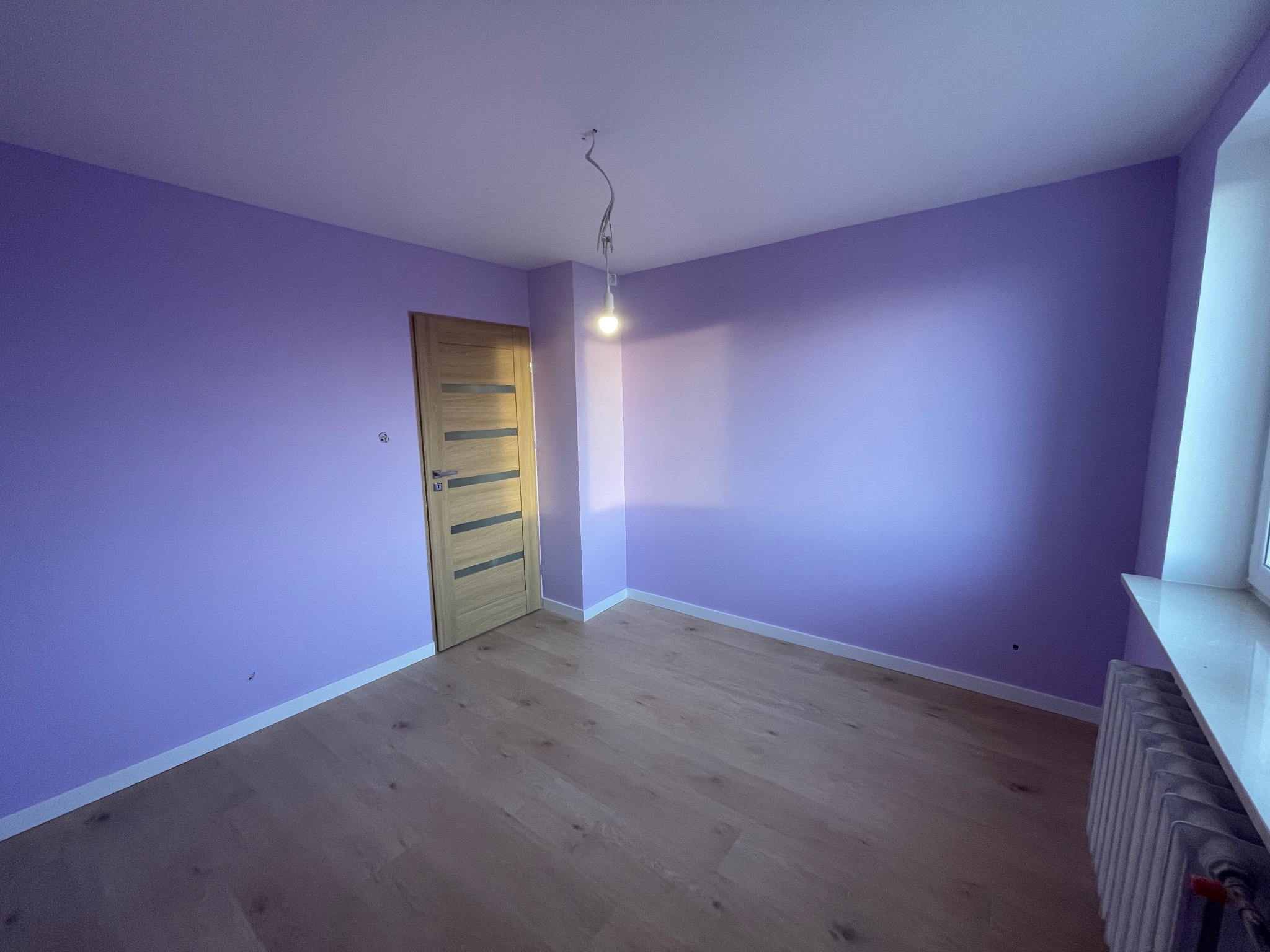 Fioletowy pokój po malowaniu: drewniana podłoga, drzwi z poziomymi paskami, białe listwy przypodłogowe i grzejnik pod oknem. Wisząca żarówka.