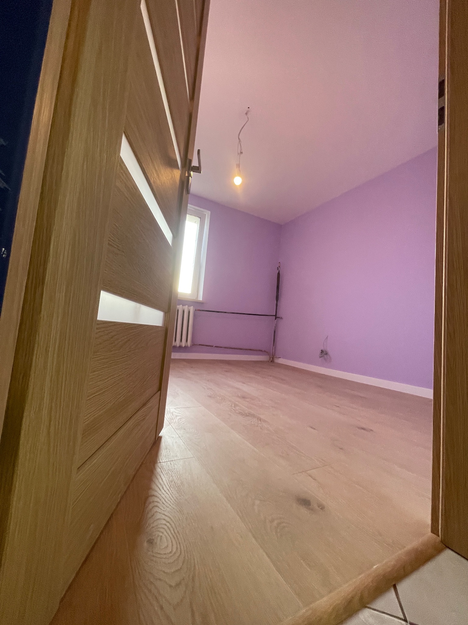 Otwarty widok na pokój z fioletowymi ścianami i jasną drewnianą podłogą, przez nowoczesne drzwi z elementami szkła. Widoczny grzejnik i surowa instalacja oświetleniowa.