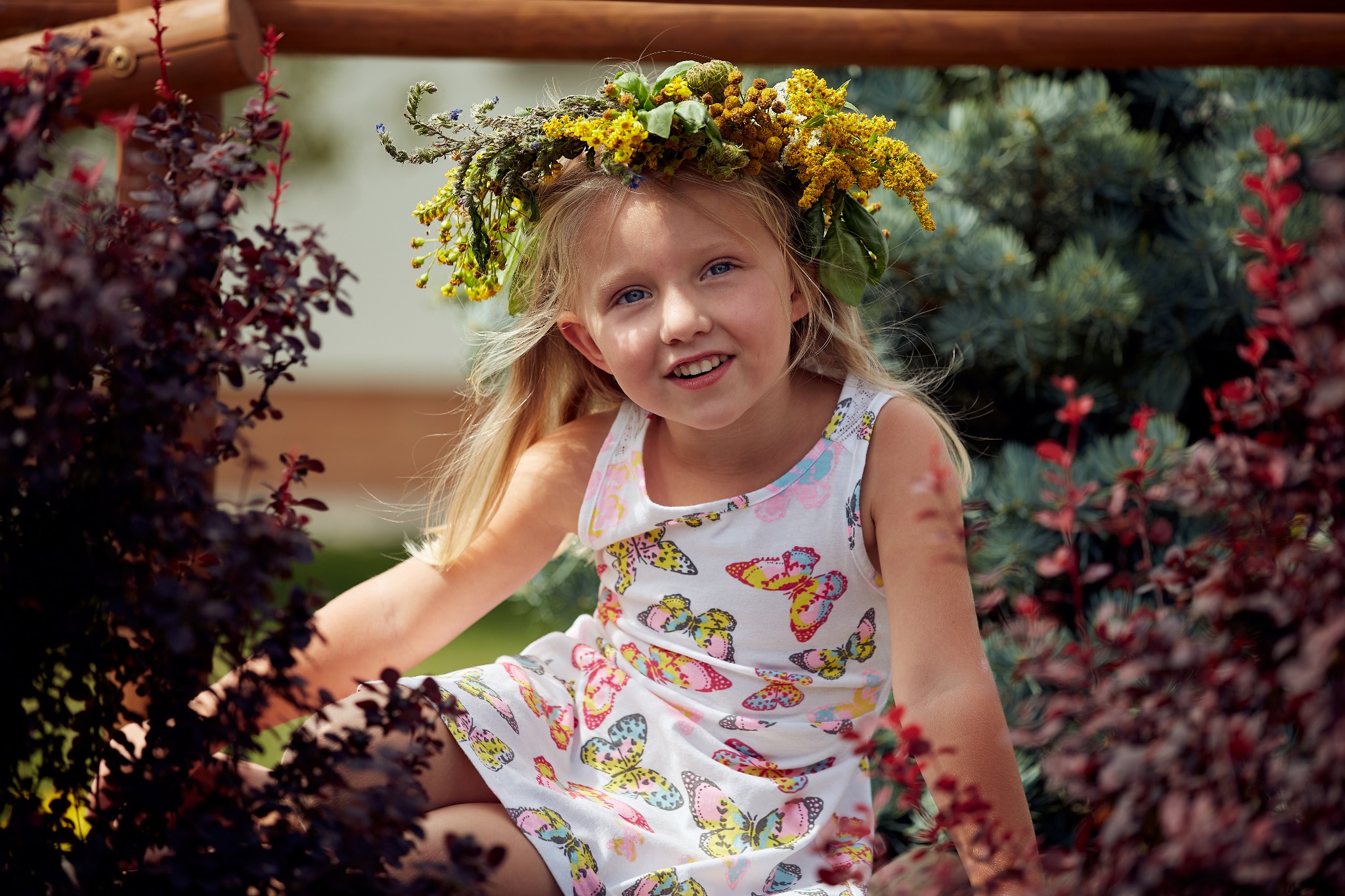 Uśmiechnięta blond dziewczynka w wianku z żółtych kwiatów, ubrana w sukienkę w motyle, siedzi w otoczeniu krzewów o bordowych liściach, w słoneczny dzień.