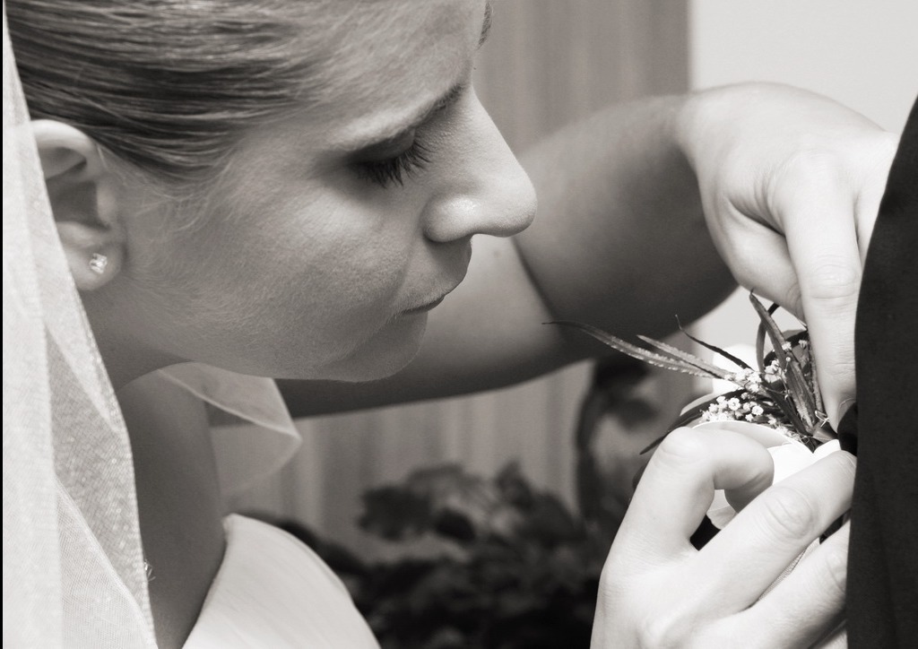 Czarno-białe zbliżenie panny młodej w welonie, poprawiającej butonierkę pana młodego. Delikatne światło, skupienie na detalu, intymna chwila przed ceremonią.