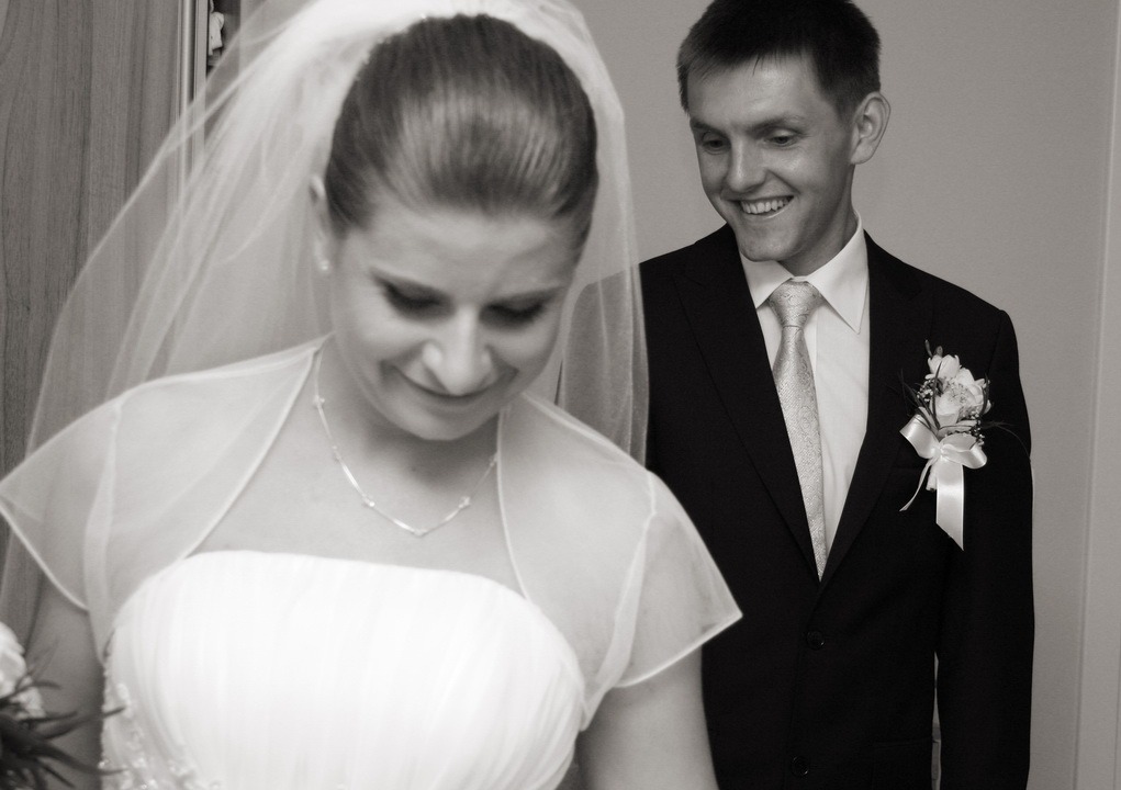 Czarno-białe zdjęcie uśmiechniętej panny młodej w sukni i welonie, spoglądającej w dół, obok uśmiechniętego pana młodego w garniturze z butonierką.