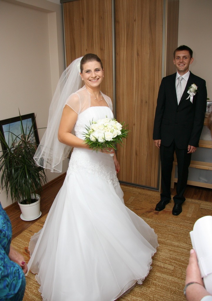 Uśmiechnięta panna młoda w białej sukni z bukietem białych róż, obok pan młody w czarnym garniturze, na tle drewnianej szafy przesuwnej i zielonej rośliny doniczkowej.