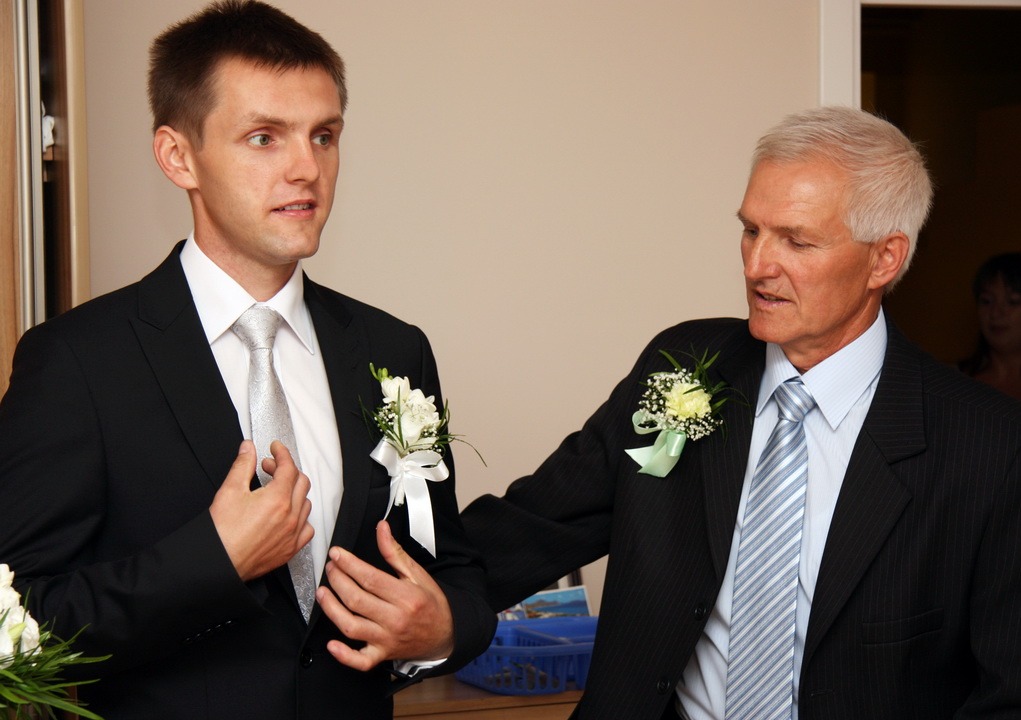 Dwóch elegancko ubranych mężczyzn w garniturach z butonierkami, prawdopodobnie podczas uroczystości ślubnej. Jeden z nich poprawia krawat.