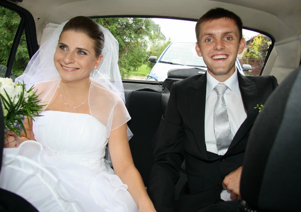 Uśmiechnięta para młoda w samochodzie: panna młoda w białej sukni z bukietem, pan młody w czarnym garniturze i srebrnym krawacie. W tle widoczny fragment niebieskiego auta.