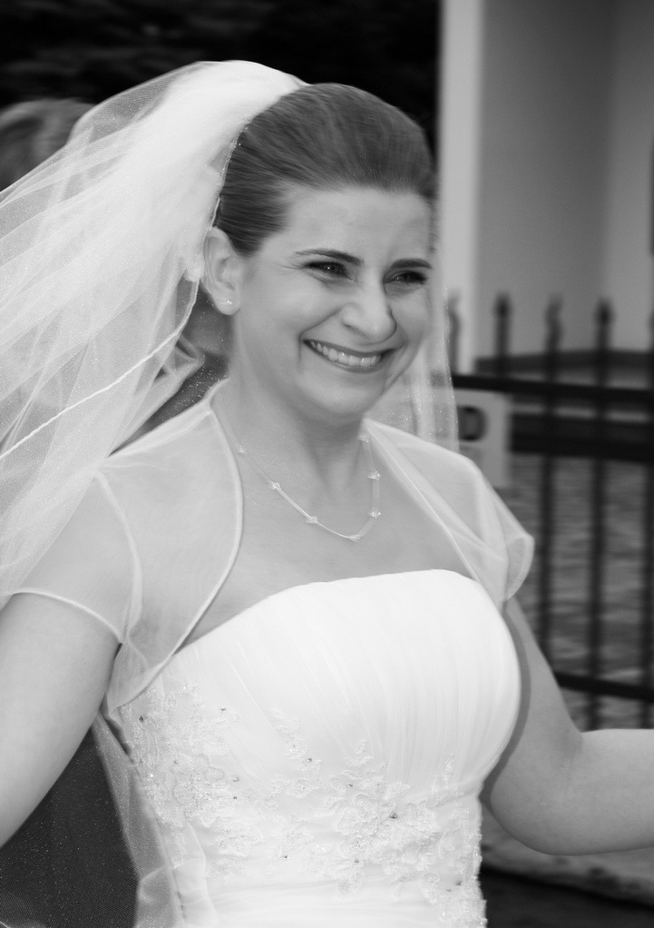 Uśmiechnięta panna młoda w białej sukni i welonie, czarno-białe zdjęcie z delikatnym rozmyciem tła, ukazujące detale sukni i biżuterii.