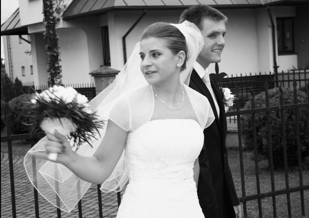 Czarno-białe zdjęcie pary młodej: panna młoda w sukni z bukietem, obok pan młody w garniturze, oboje uśmiechnięci, na tle budynku i ogrodzenia.