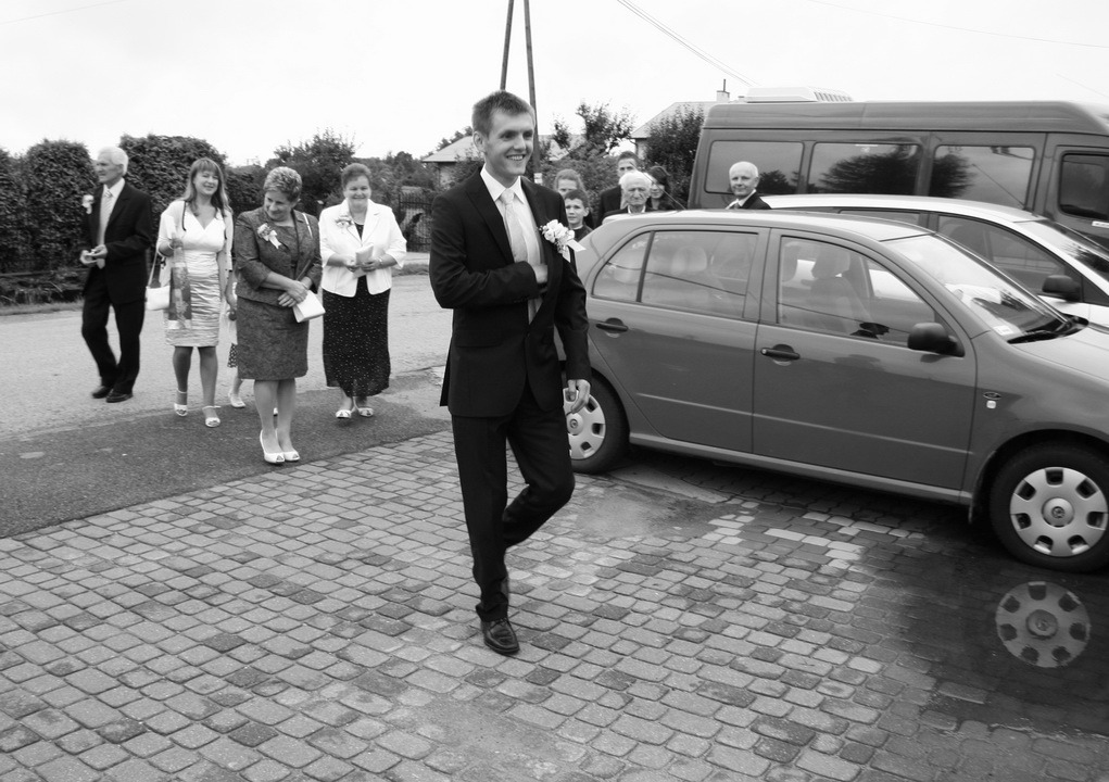 Czarno-białe zdjęcie uśmiechniętego pana młodego w garniturze, idącego po brukowanej drodze, z gośćmi weselnymi i samochodami w tle. Ujęcie z niskiej perspektywy.