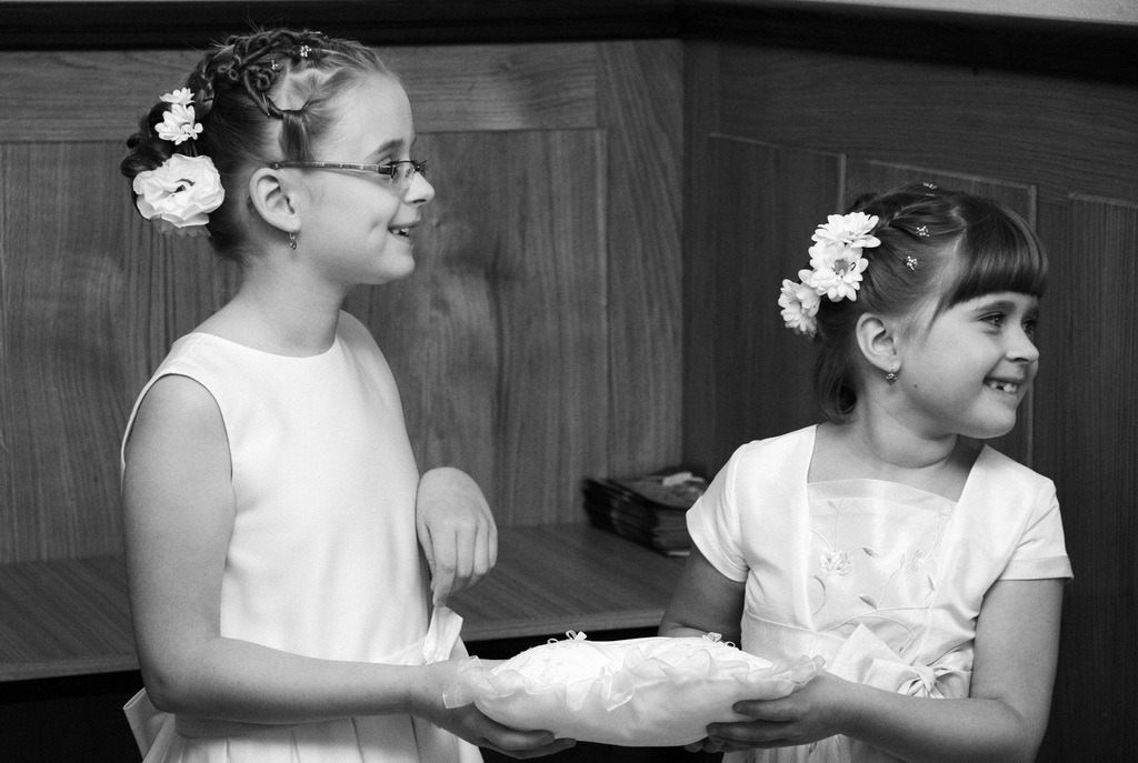 Dwie uśmiechnięte dziewczynki w białych sukienkach, z kwiatami we włosach, trzymają poduszkę na obrączki. Zdjęcie czarno-białe, z delikatnym oświetleniem.
