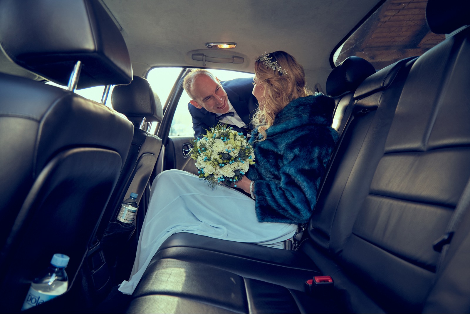 Uśmiechnięty pan młody w samochodzie wręcza bukiet pannie młodej w błękitnym futrze i sukni. W tle widoczne butelki wody. Romantyczna chwila przed ślubem.