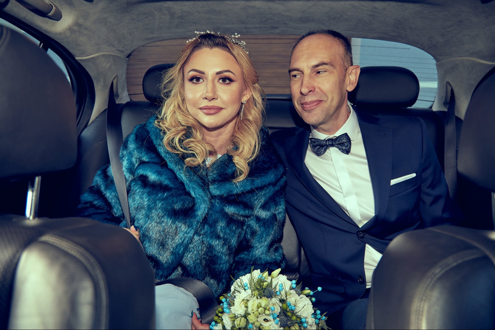 Para młoda w samochodzie: Panna młoda w turkusowym futrze i tiarze, Pan młody w granatowym garniturze z muchą. Bukiet ślubny w dłoniach panny młodej. Uśmiechnięci, w drodze na ceremonię.