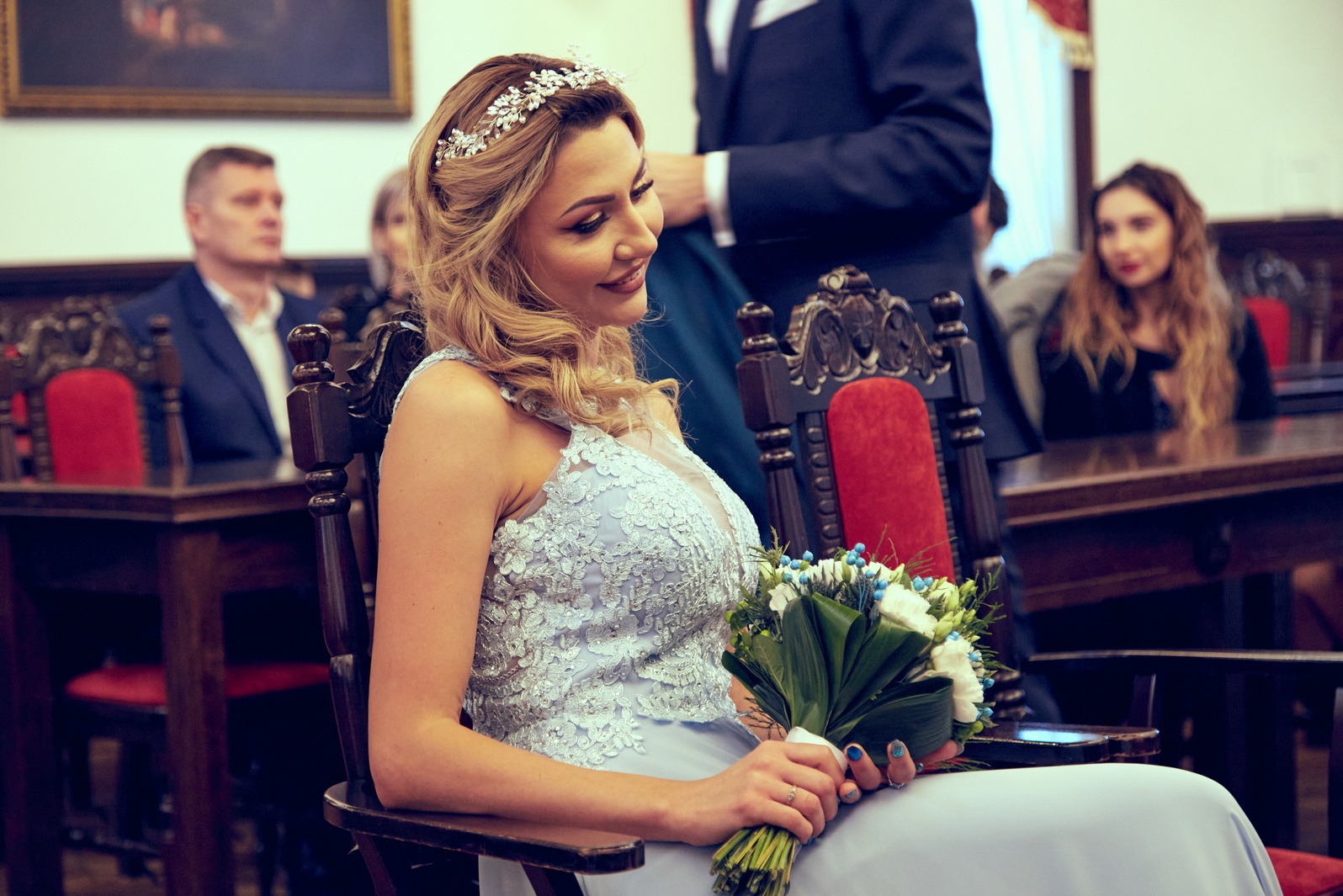 Panna młoda w koronie i błękitnej sukni siedzi na krześle, trzyma bukiet. W tle goście i klasyczne wnętrze sali ślubów. Uśmiechnięta, elegancka stylizacja.
