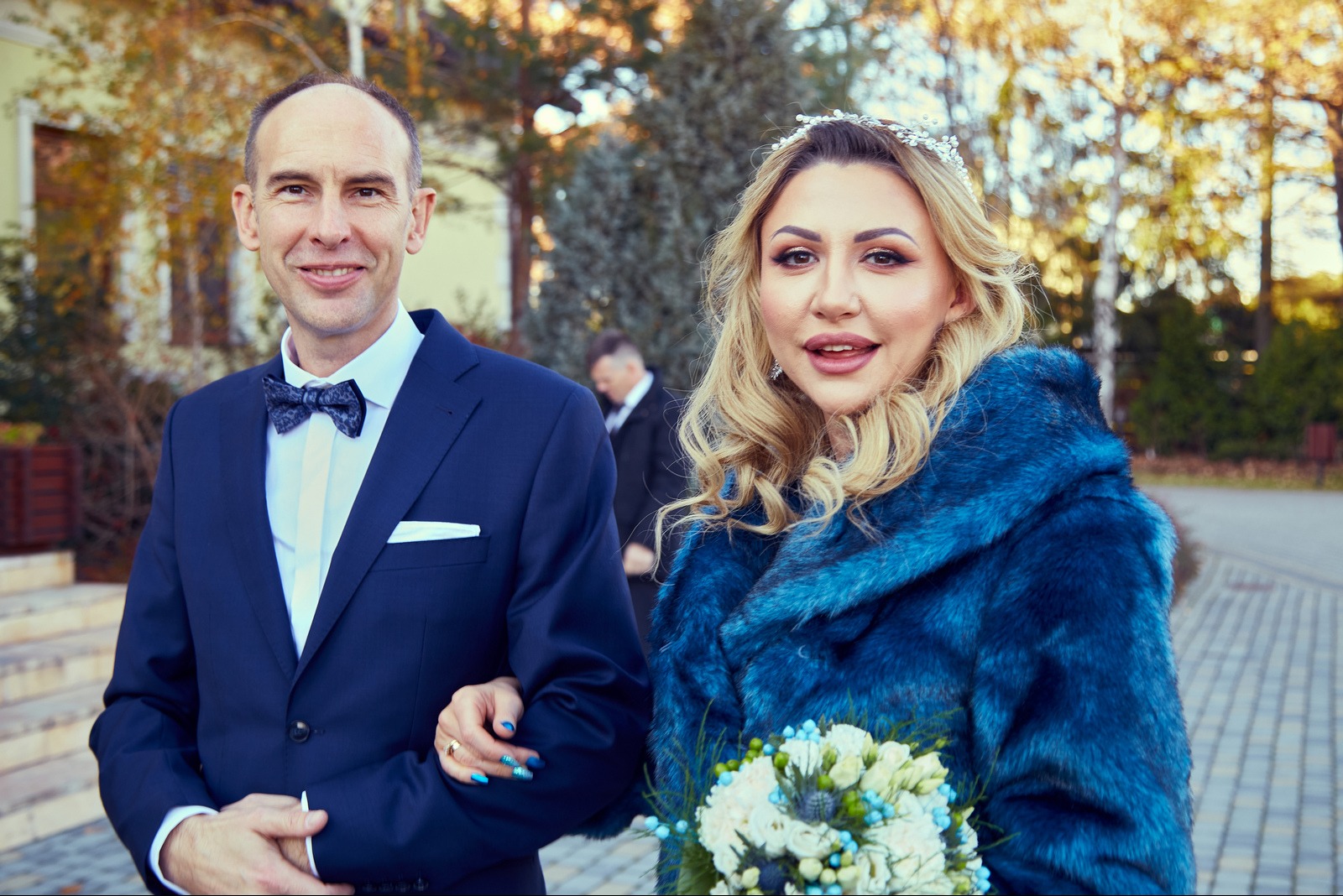 Uśmiechnięta para młoda: mężczyzna w granatowym garniturze i kobieta w błękitnym futrze z bukietem, pozują na tle jesiennego krajobrazu. Ujęcie z bliska, naturalne światło.