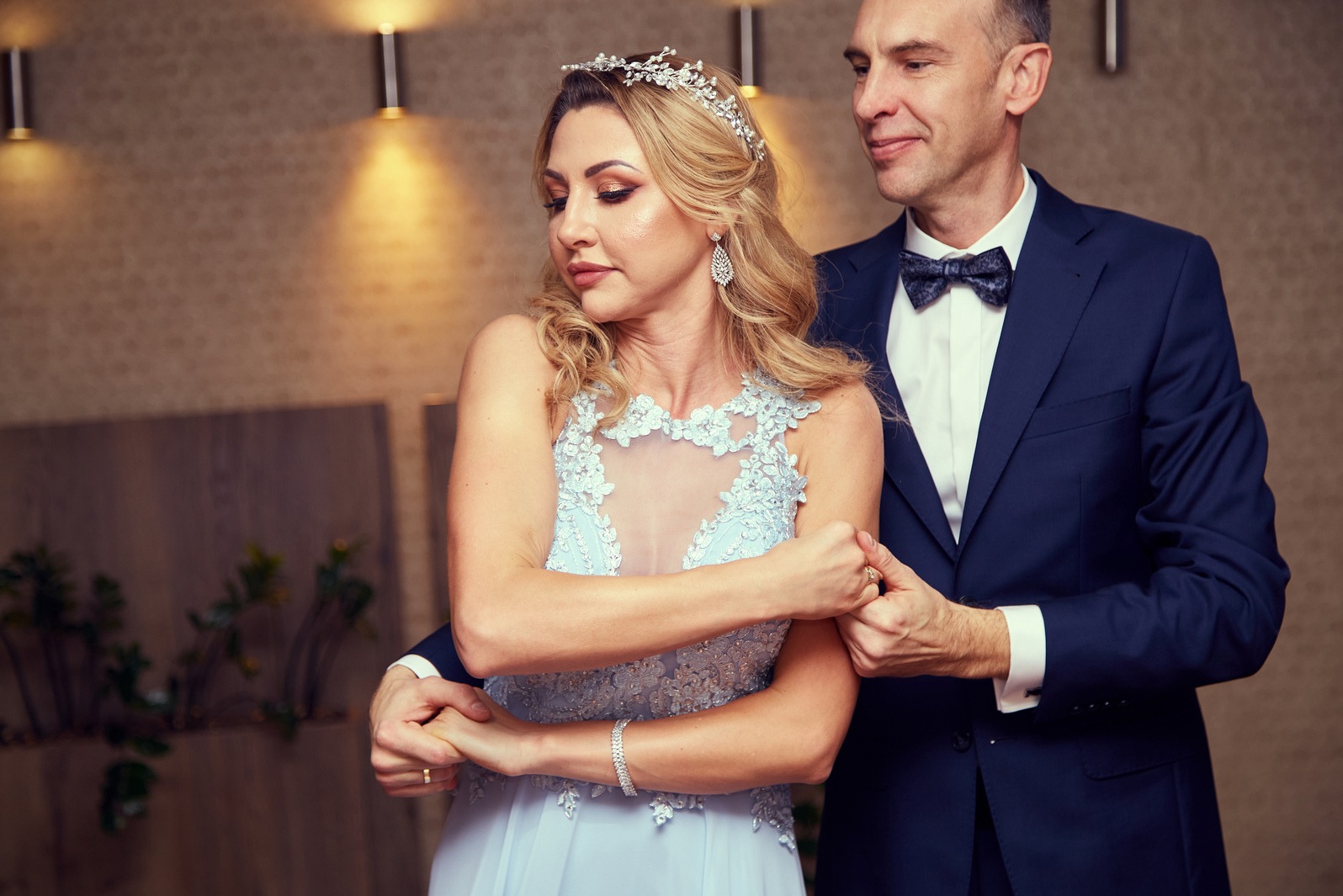 Elegancka para: blond kobieta w błękitnej sukni i diademie, obejmowana przez mężczyznę w granatowym garniturze z muchą, pozuje na tle dekoracyjnej ściany.