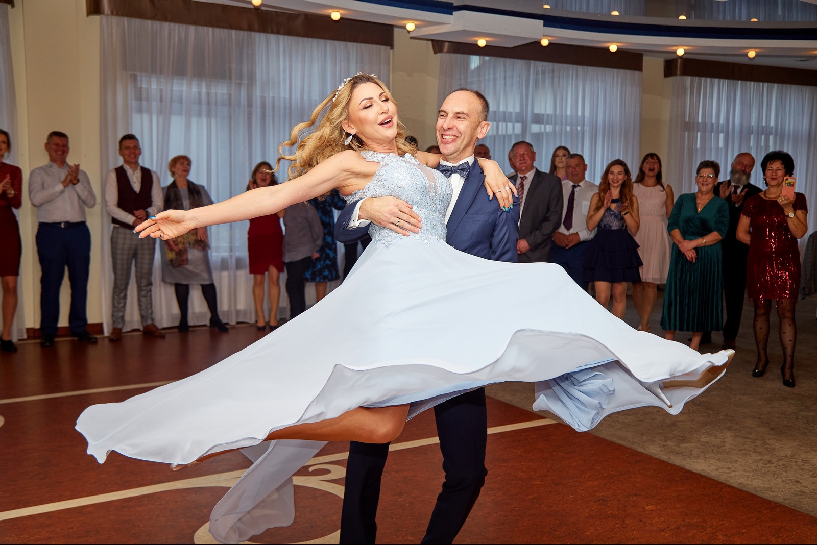 Radosny taniec pary młodej w Sokołowie Małopolskim. Pan młody trzyma pannę młodą w błękitnej sukni, goście weselni w tle klaszczą.