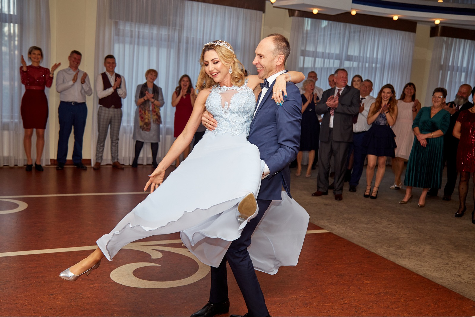 Uśmiechnięty pan młody w granatowym garniturze trzyma na rękach pannę młodą w jasnej sukni podczas pierwszego tańca, w tle goście weselni.