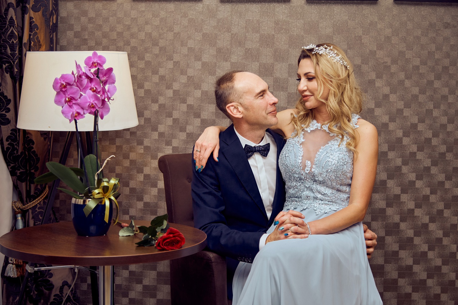 Elegancka sesja narzeczeńska: para w objęciach, ona w błękitnej sukni i diademie, on w smokingu, w tle wzorzysta ściana, lampa z orchideami i róża na stoliku.