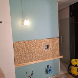 Alexey Martsinovskiy - Ściana w łazience w trakcie remontu. Częściowo ułożona mozaika, widoczne otwory na instalacje, płyta gipsowa i lampa wisząca. Prace wykończeniowe w toku.