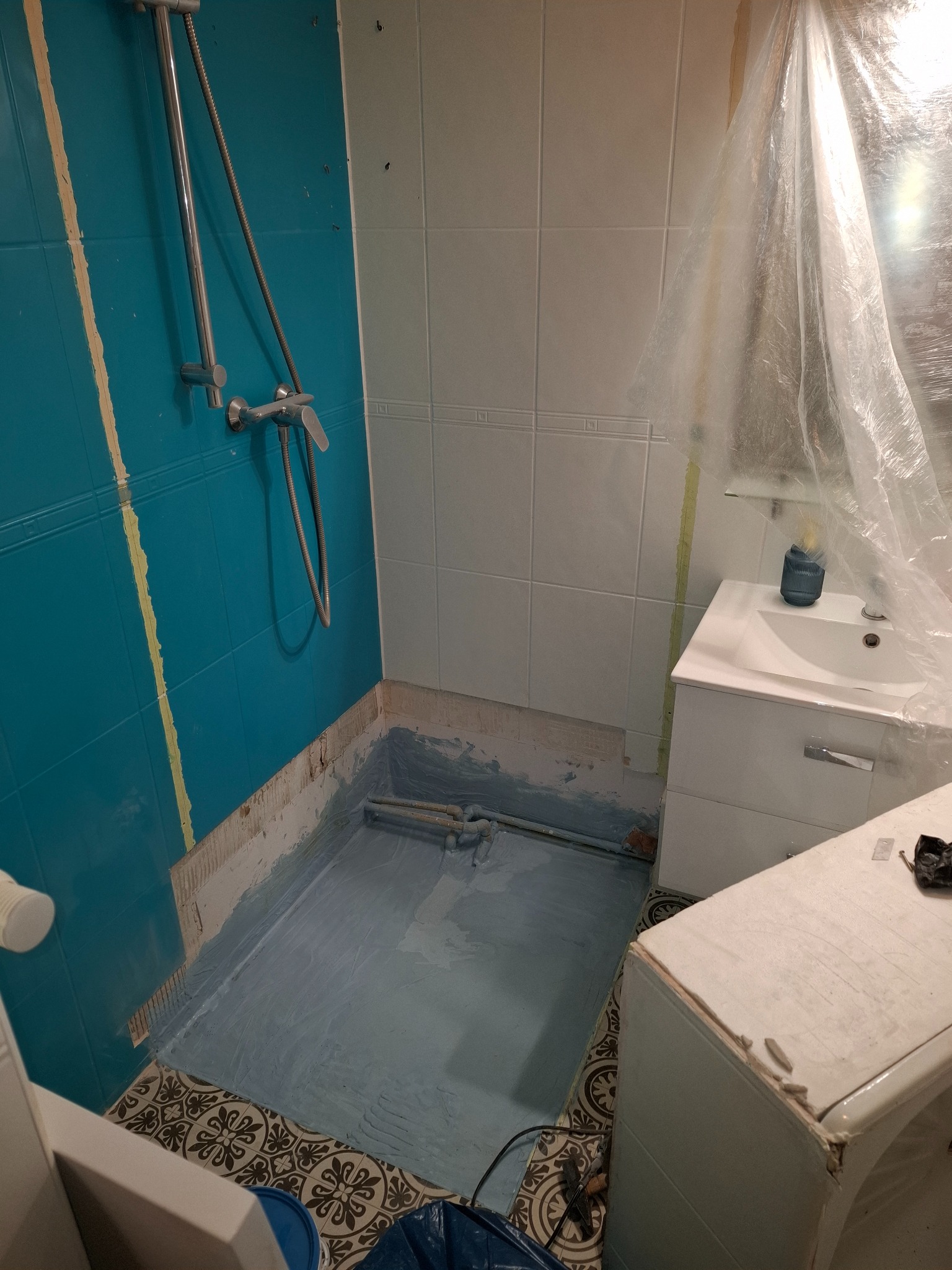 Łazienka w trakcie remontu: zabezpieczone elementy, niebieska hydroizolacja brodzika, częściowo usunięte płytki, widoczna armatura i fragment pralki.