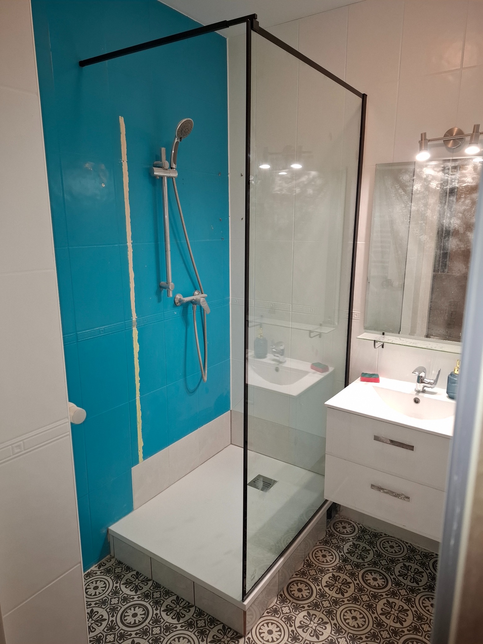 Narożna kabina prysznicowa z czarną ramą i turkusową ścianą w łazience z wzorzystą podłogą. Widoczna umywalka z szafką i lustro.