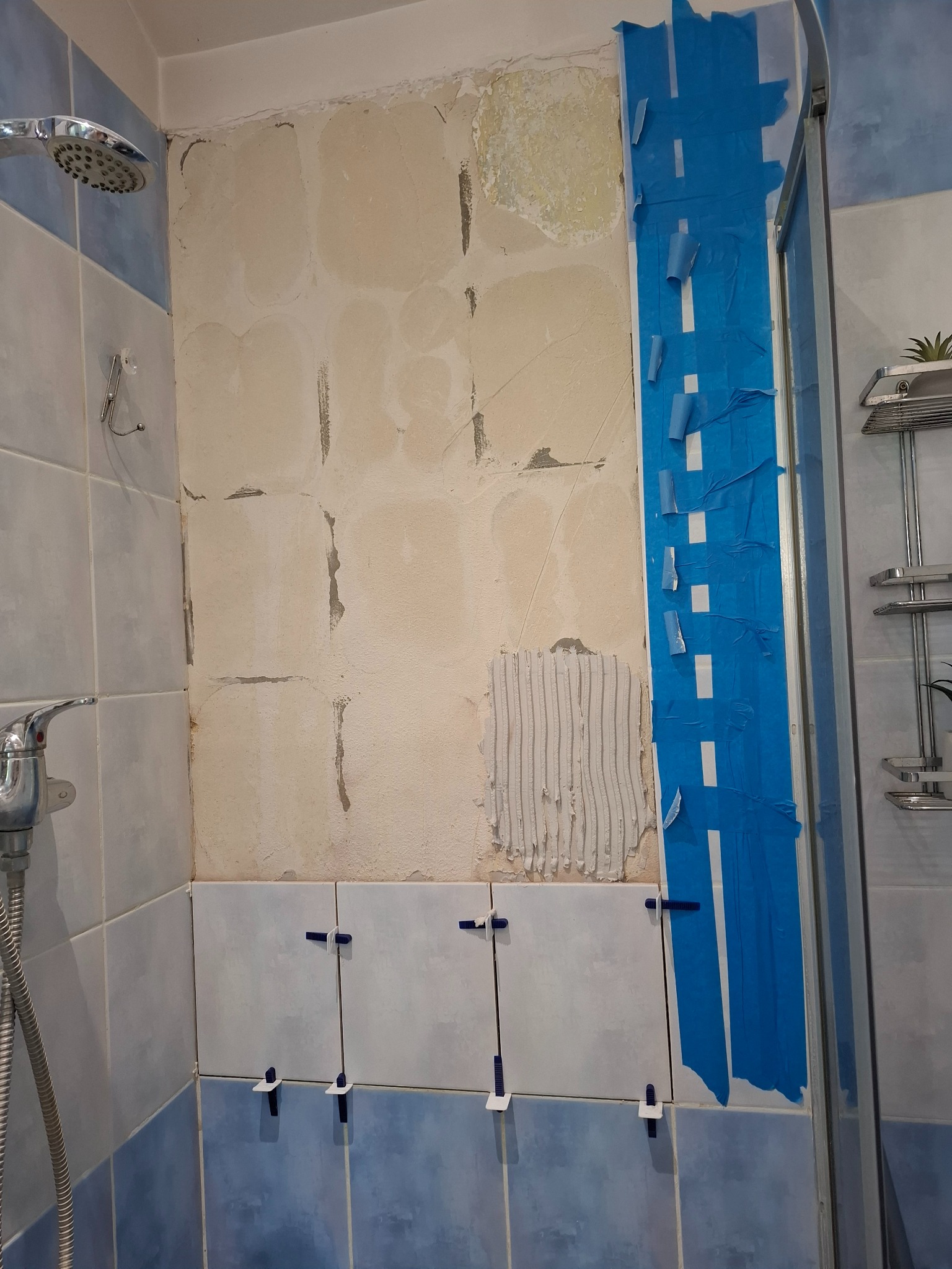 Fragment łazienki w trakcie remontu: częściowo usunięte stare płytki, świeżo nałożony klej, nowe płytki z krzyżykami dystansowymi, niebieska taśma malarska.