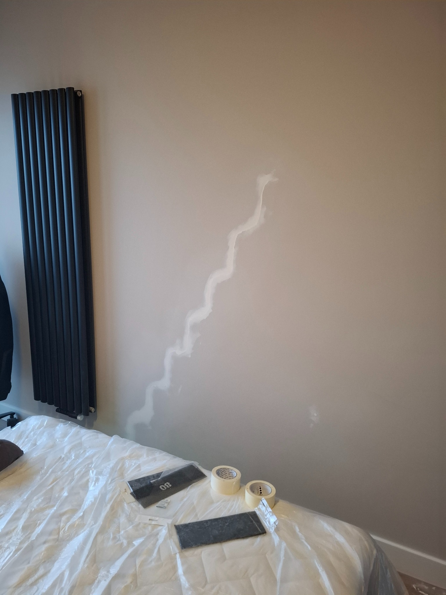 Szpachlowanie ściany w zygzak w Zielonce. Widoczny grzejnik, zakryte łóżko folią, szpachelki i taśma malarska. Przygotowanie do malowania, etap prac wykończeniowych.