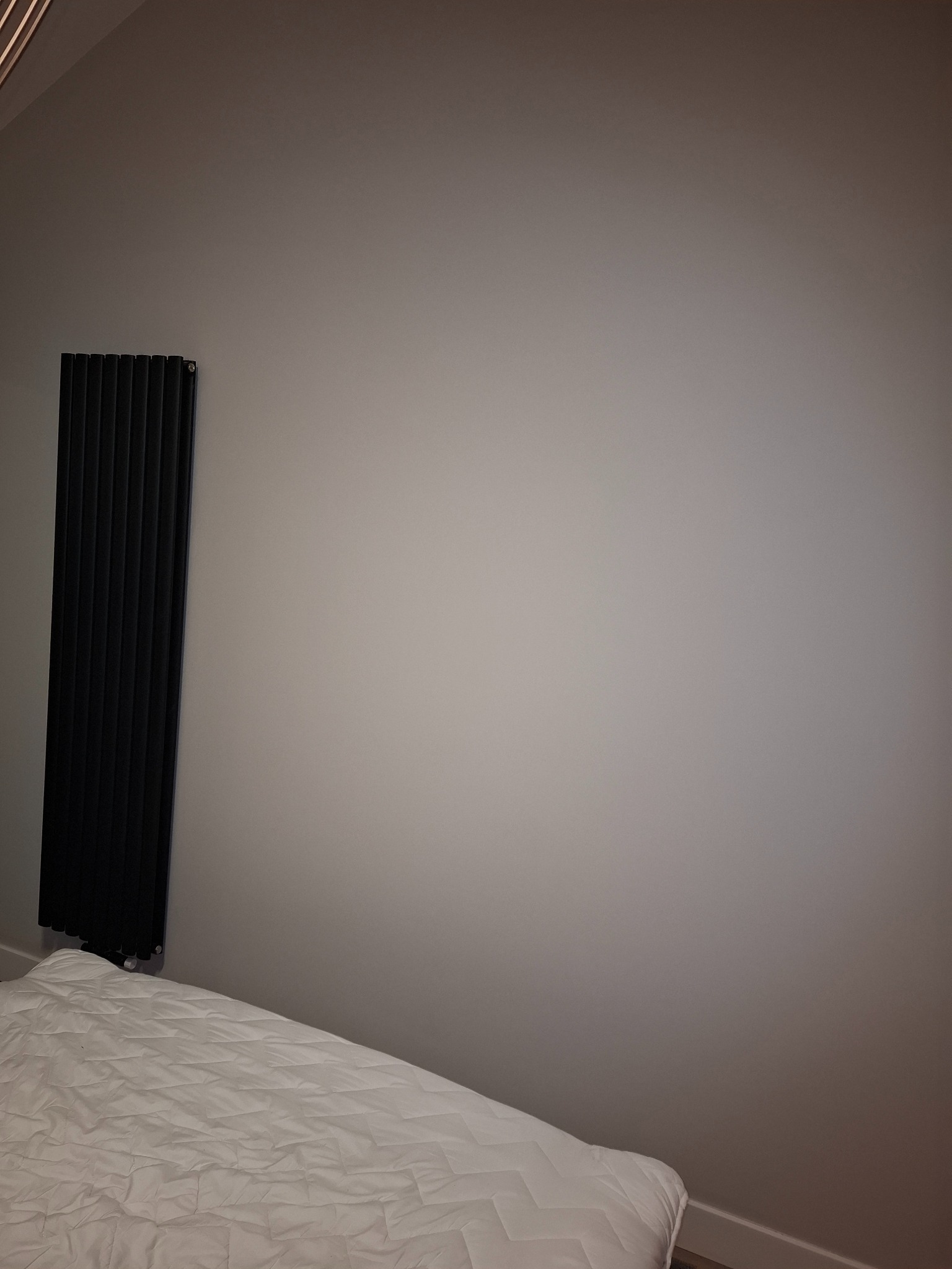 Czarny, pionowy grzejnik panelowy zamontowany na szarej ścianie w pokoju, widoczny fragment białej kołdry. Minimalistyczny design, oświetlenie z góry.