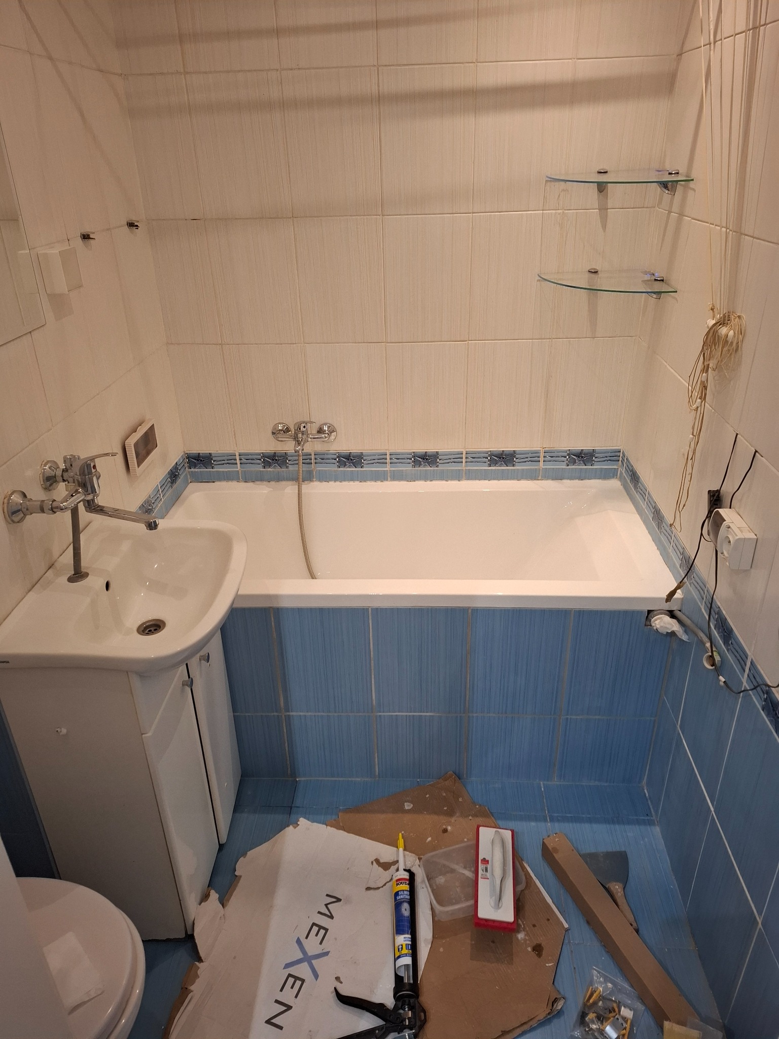 Łazienka w trakcie remontu: biała wanna obudowana niebieskimi płytkami, umywalka z szafką, narzędzia i materiały budowlane na podłodze. Widoczne szklane półki.