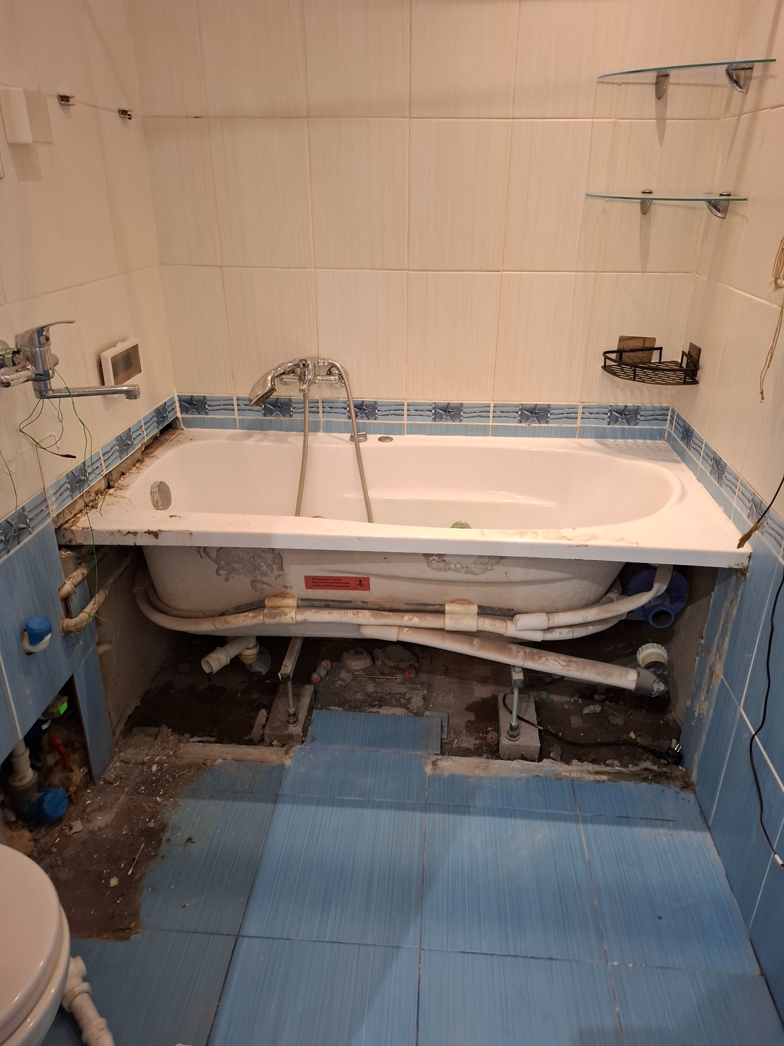 Demontaż wanny podczas remontu łazienki. Widoczne rury, płytki w odcieniach bieli i błękitu, półki szklane. Ujęcie z góry, ukazujące etap prac.