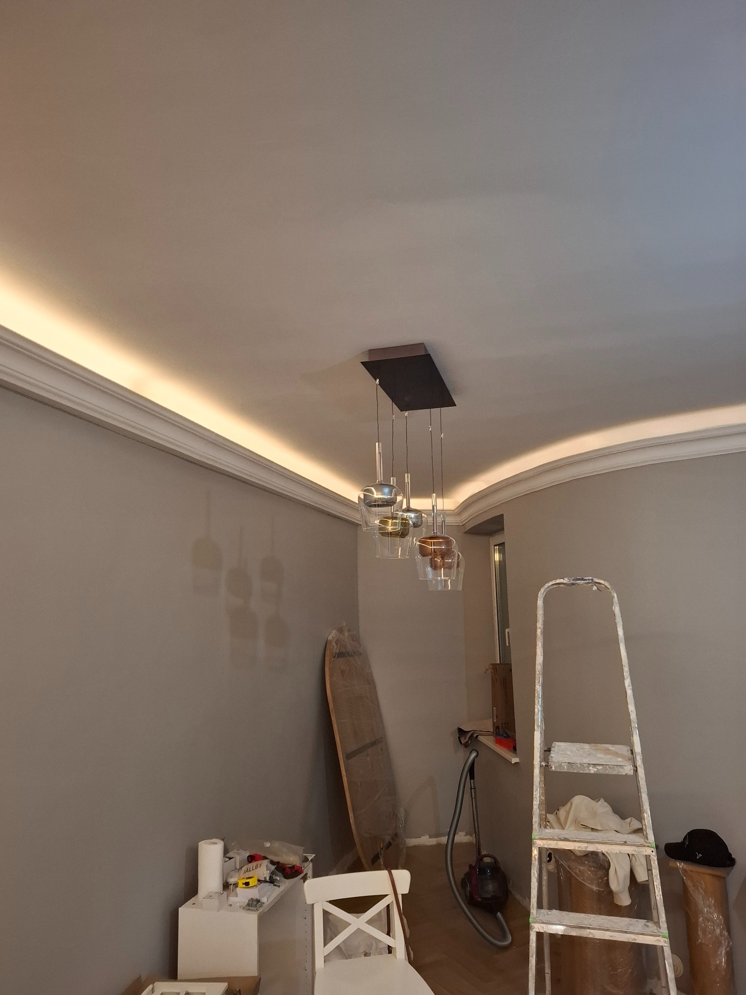 Nowoczesna lampa wisząca z kloszami w odcieniach szarości i złota, zamontowana w pokoju z listwami oświetleniowymi LED. Widoczna drabina i akcesoria malarskie.