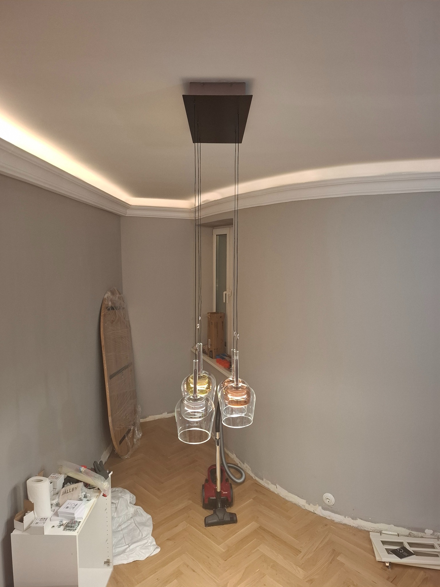 Nowoczesna lampa wisząca z trzema szklanymi kloszami, zamontowana w szarym pokoju z listwami LED pod sufitem. Widoczne elementy wykończeniowe i odkurzacz.