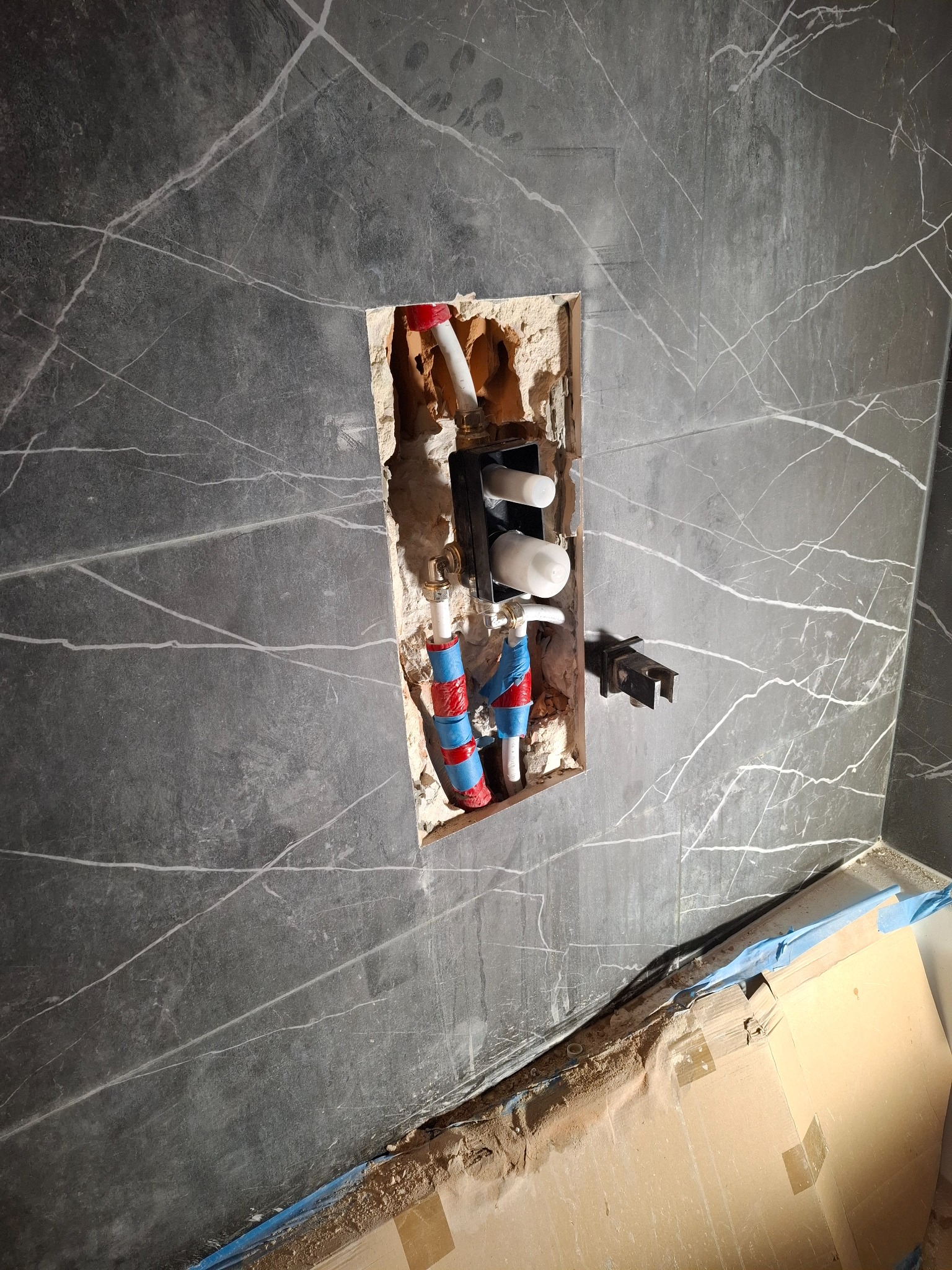 Szare płytki imitujące marmur z wycięciem na instalację wodną z rurami w kolorze czerwono-niebieskim. Widoczne elementy montażowe i fragment zabezpieczonej podłogi.