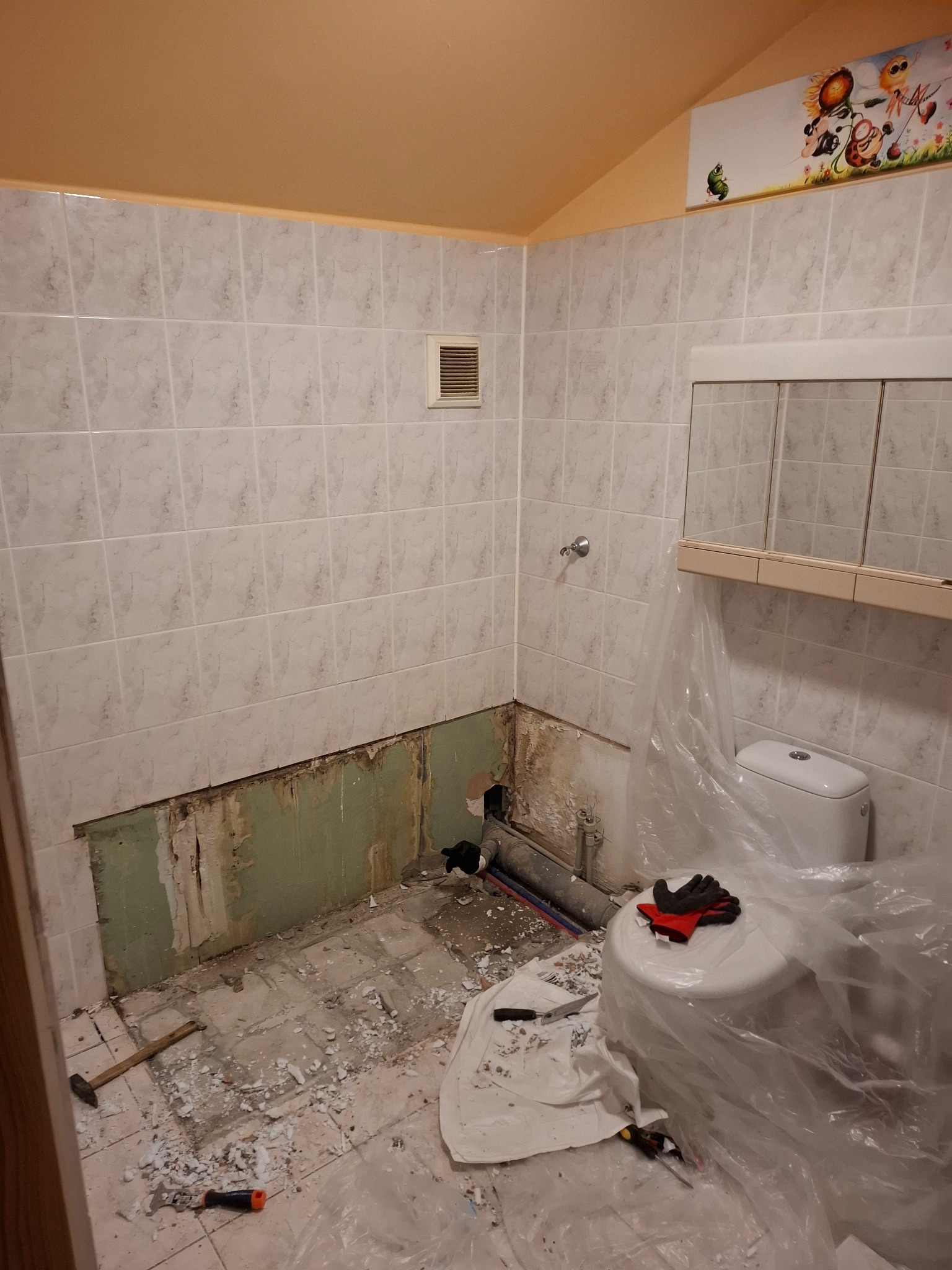 Demontaż płytek w łazience z widocznymi narzędziami i zabezpieczonym WC folią. Fragment ściany bez płytek, odsłonięta instalacja. Górna część ściany wykończona płytkami.