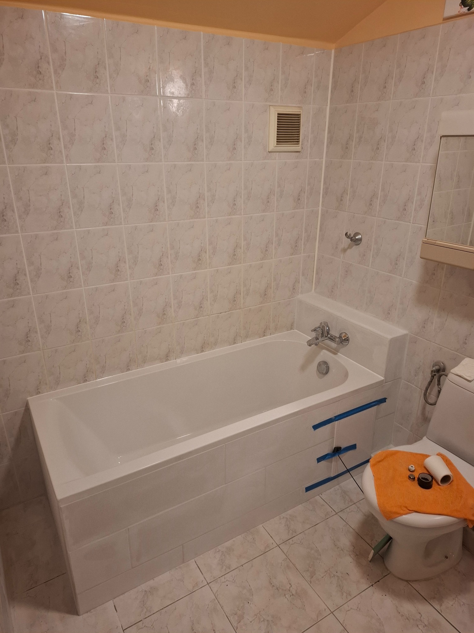 Wykończona łazienka z białą wanną obudowaną płytkami, widoczna toaleta z pomarańczowym ręcznikiem i narzędziami. Ściany wyłożone jasnymi płytkami imitującymi marmur.