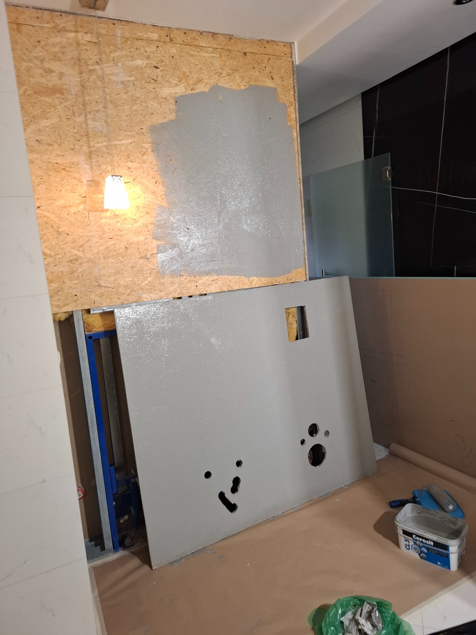 Łazienka w trakcie remontu: płyta OSB z szarą farbą, zabudowa stelaża WC, widoczny stelaż, puszka z farbą, wałek malarski. Prace wykończeniowe w toku.