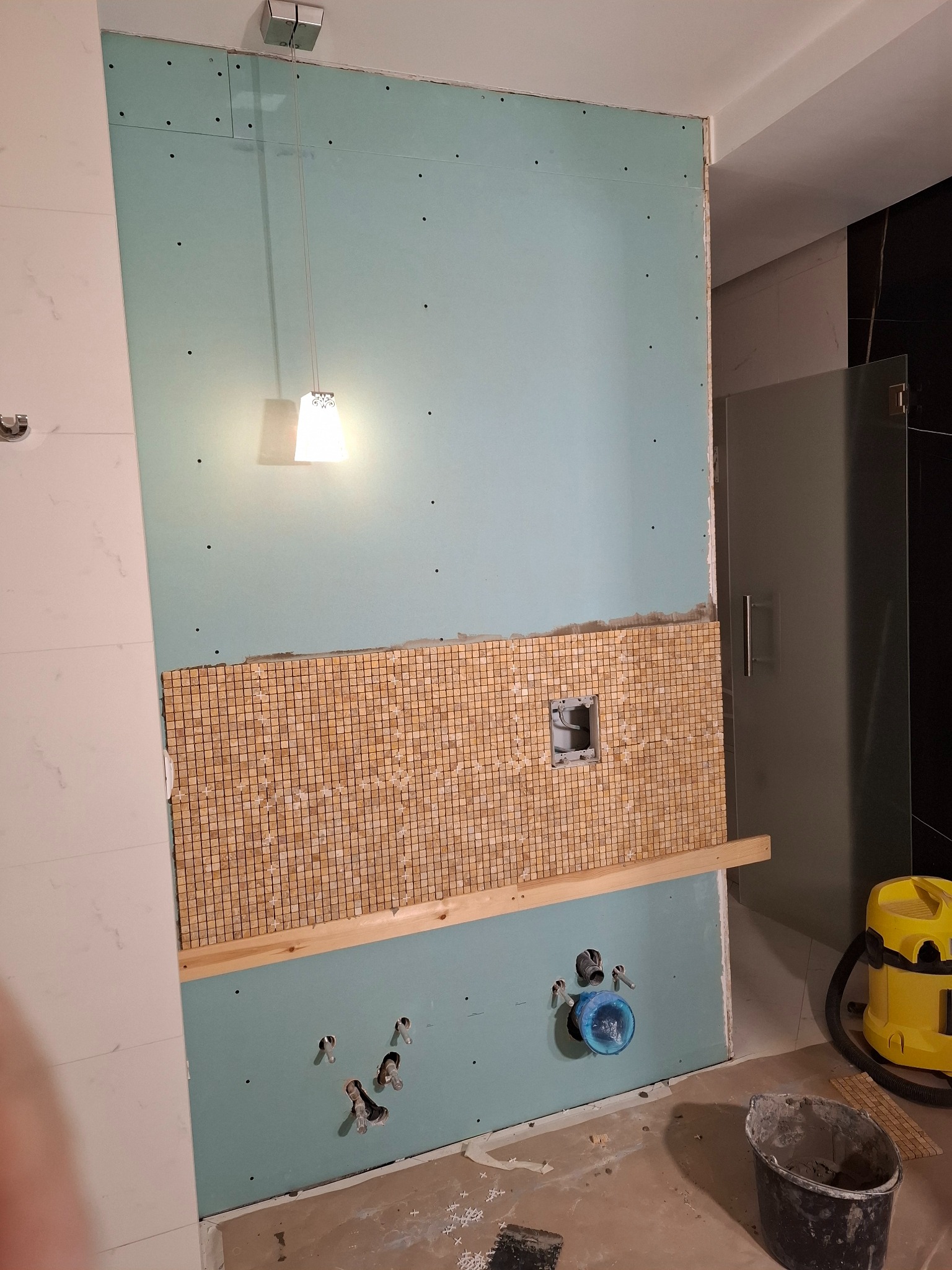 Ściana w łazience w trakcie remontu. Częściowo ułożona mozaika, widoczne otwory na instalacje, płyta gipsowa i lampa wisząca. Prace wykończeniowe w toku.