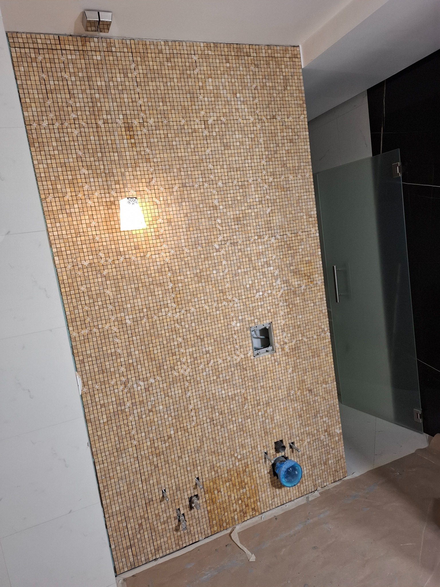 Ściana łazienki wyłożona mozaiką w odcieniach beżu i brązu, z widocznymi otworami na armaturę i fragmentem kabiny prysznicowej. U góry zamontowane oświetlenie.