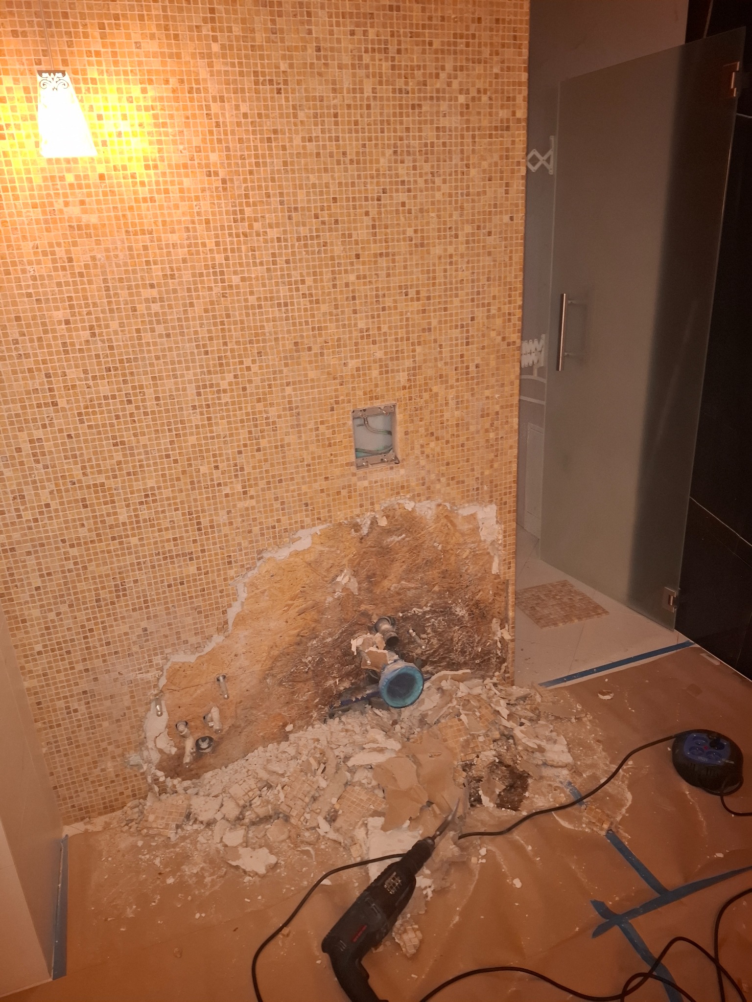 Demontaż mozaiki w łazience z widocznymi rurami i gruzem, w tle kabina prysznicowa. Użyto wiertarki udarowej. Etap prac przygotowawczych do remontu.