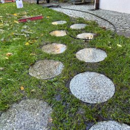 Sebastian Knapik - Mokre, okrągłe płyty chodnikowe z szarego kamienia, ułożone w trawie, po umyciu. W tle widać myjkę ciśnieniową i szczotkę.