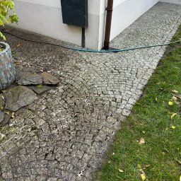 Sebastian Knapik - Mokry fragment kostki brukowej po czyszczeniu w Ksawerowie, widoczny wzdłuż ściany budynku i trawnika. W tle rura spustowa i skrzynka na ścianie.