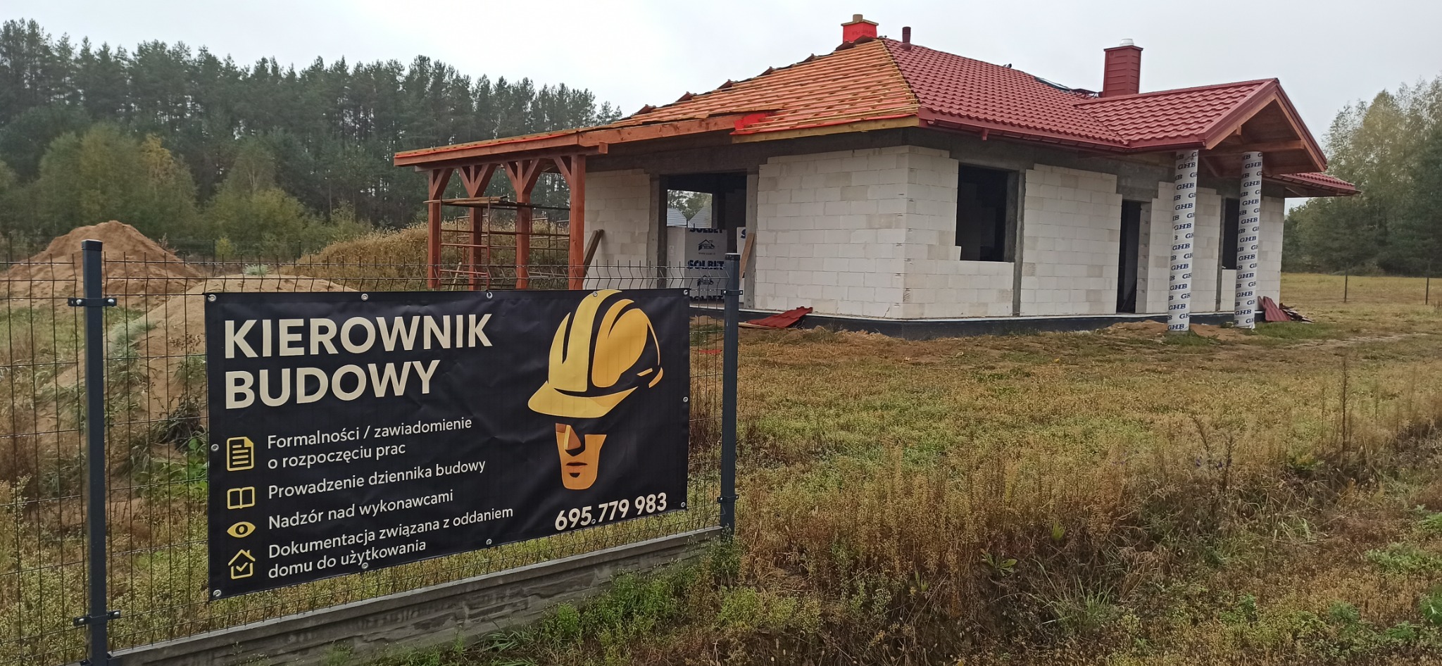 Budowa domu jednorodzinnego w trakcie realizacji, widoczny budynek w stanie surowym otwartym i baner informacyjny z numerem telefonu.