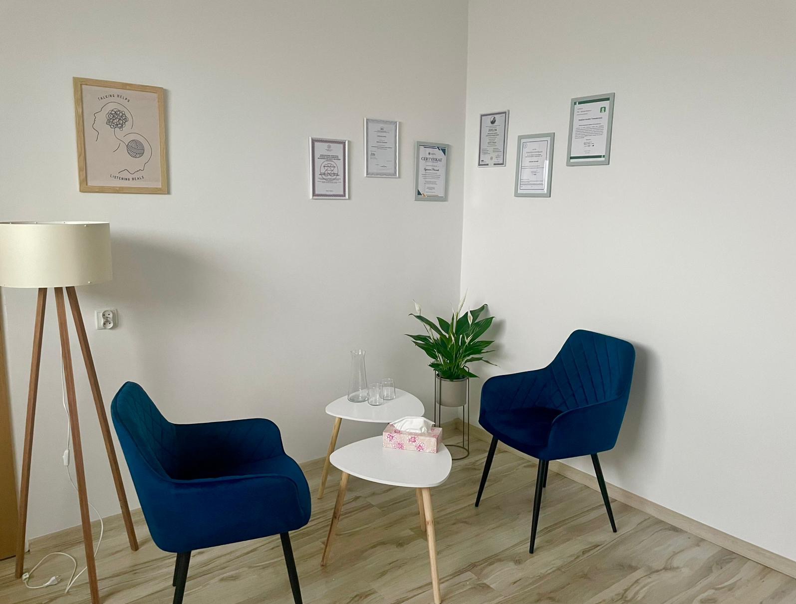 Jasne wnętrze gabinetu z dwoma granatowymi fotelami, stolikami, lampą i dyplomami na ścianie. Spokojna aranżacja przestrzeni do rozmów.