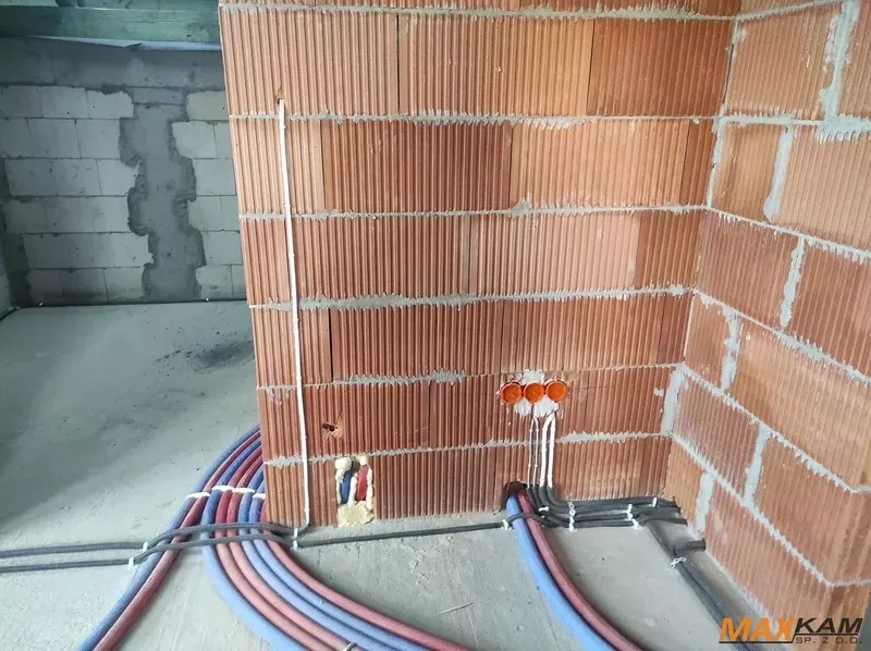 Instalacja elektryczna i wodna w surowym wnętrzu: pomarańczowe puszki podtynkowe, białe przewody, kolorowe rury w peszlach na podłodze, ściany z czerwonej cegły dziurawki.