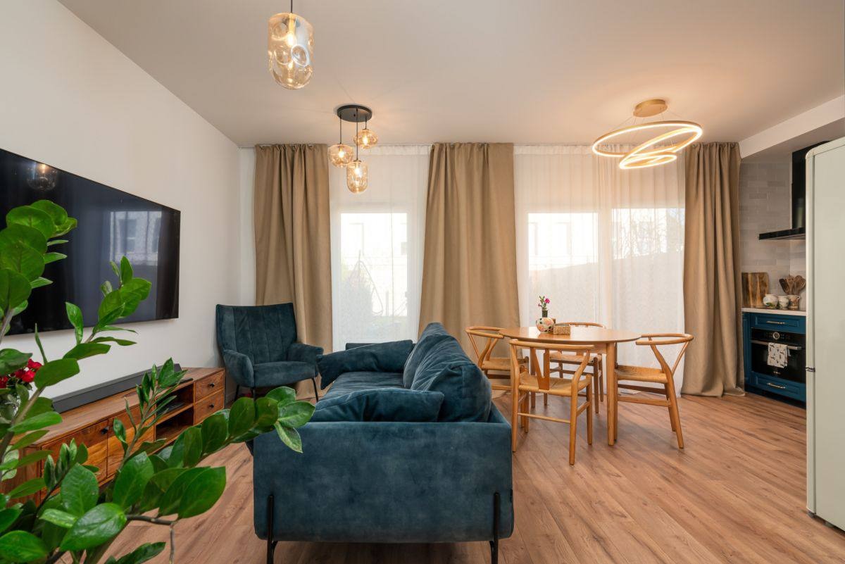 Nowoczesny salon z drewnianą podłogą, turkusową sofą i fotelami. Okrągły stół z krzesłami, designerskie lampy wiszące. Widok na kuchnię w tle.