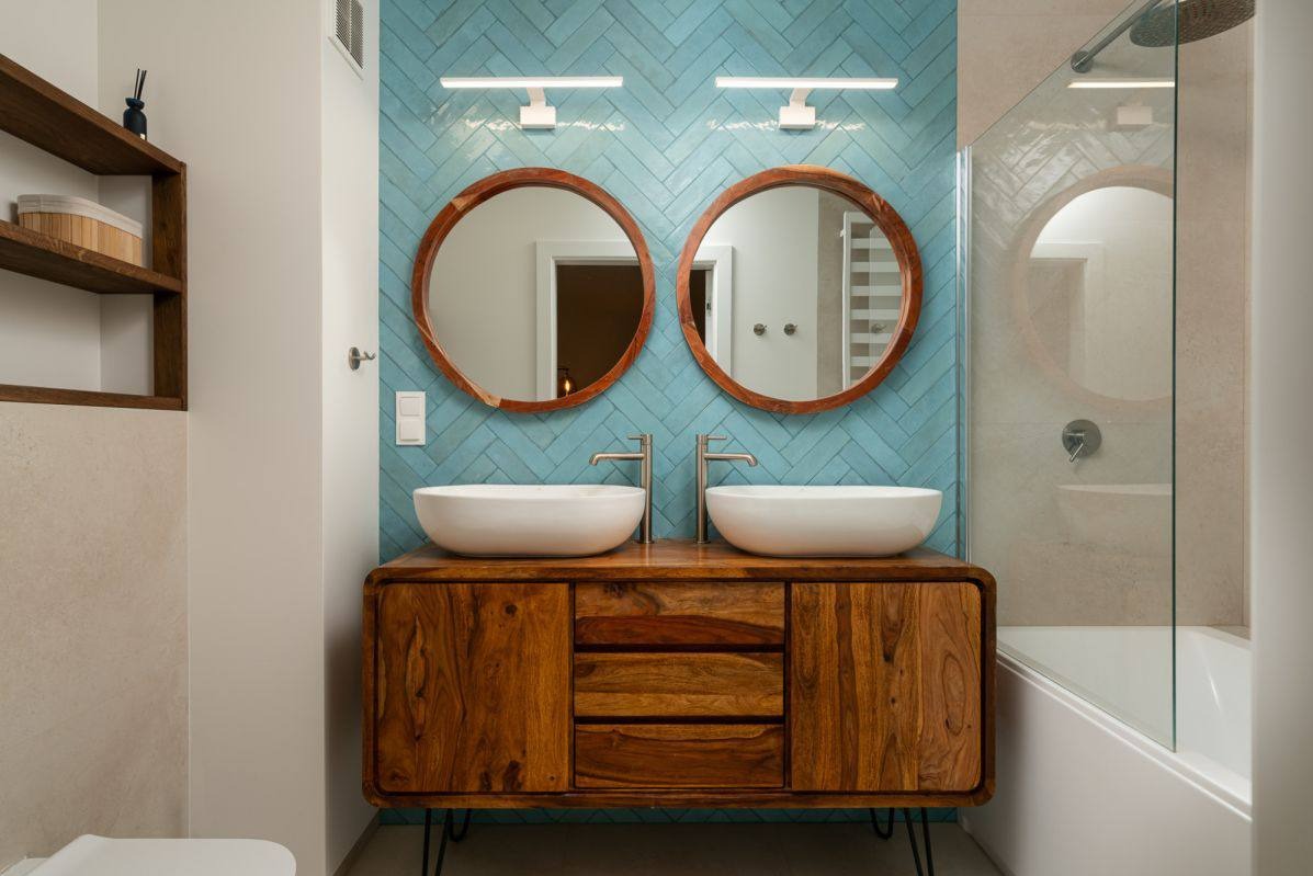 Nowoczesna łazienka z drewnianą szafką pod umywalki, okrągłymi lustrami i turkusową ścianą z płytek ułożonych w jodełkę. Widoczna kabina prysznicowa.